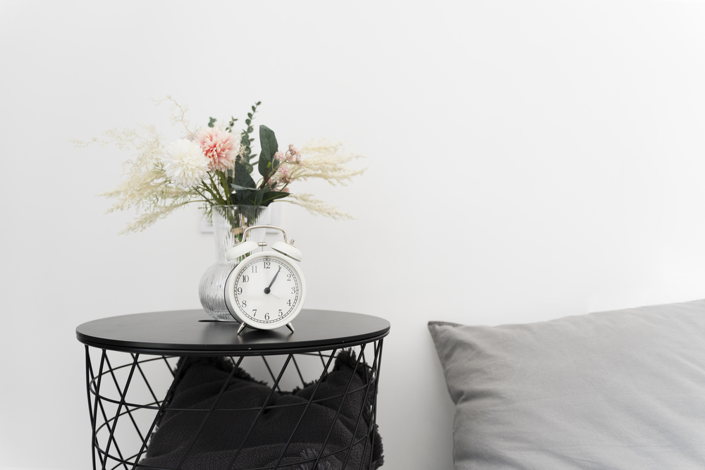 Mesita de noche moderna y sencilla decorada con flores y un reloj clásico, que aporta frescura y estilo funcional al dormitorio.